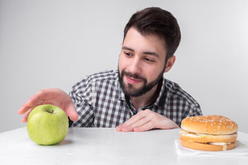 Bearded man in checkered shirt on a light background holding a hamburger and an apple. Guy makes the choice between fast and healthy food. Tasty or useful The dilemma