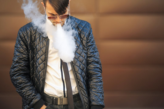Vape man. Portrait of a handsome young white guy with modern haircut in aviator sunglasses vaping an electronic cigarette opposite the futuristic urban background. Lifestyle.
