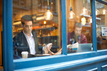 Window view of businessmen reading newspaper and using laptop in cafe