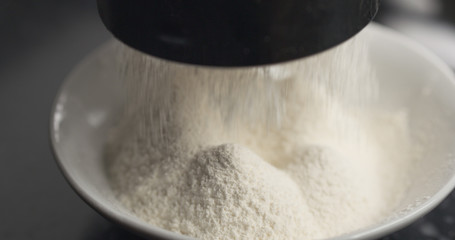 Handheld shot of sieving flour with mug sieve on plate closeup, 4k photo