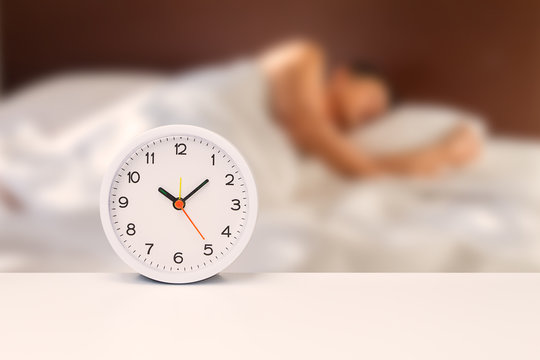 White alarm clock on white desk on blurred man sleeping on white bedding sheet, time concept
