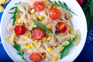 Pasta salad with tomatoes cherry, tuna, corn and arugula on blue wooden background. Top view.