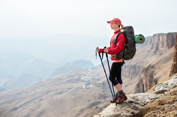 A slender girl in a cap with sticks for Nordic walking with a backpack and a folded rug for relaxation stands in the mountains against the backdrop of rocks and distant lands