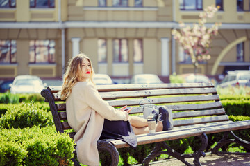 Young girl in with a cup of coffee sitting on a bench, a sunny street of the old city