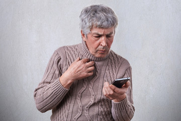 A portrait of elderly male with gray hair having shocked expression while looking into his smartphone reading message. A man looking terrible video online using mobile phone.