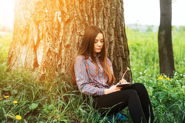 A young, pretty girl in a shirt sits near a tree on the grass on a sunny day with a notebook. - 150126416