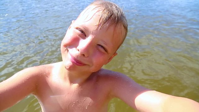 Closeup portrait of cute funny kid taking selfie on summer river beach at sparkling sunny water background. Little boy uses modern technology. Child blows kiss into camera cheerfully. Point of view.