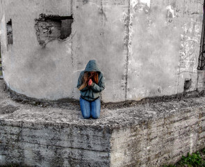 A young woman with long blonde hair in a hood tilted her head and clasped her hands in prayer. The girl prays on her knees near the gray morose wall