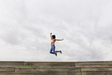 Happy young beautiful afro american woman listening to music in her mobile phone and jumping. Cloudy background. Spring or summer season. Casual