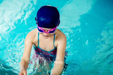 Portrait funny little girl in blue cap and pink goggles playing in swim pool. Close up