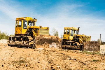 Two yellow dirty old bulldozer stand on a construction site on a sunny day against a background of forest and blue sky