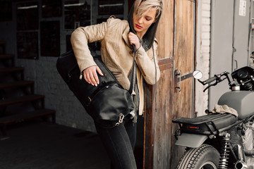 A young woman with a leather bag near the garage. A woman in a leather jacket. Motorcycle near the...