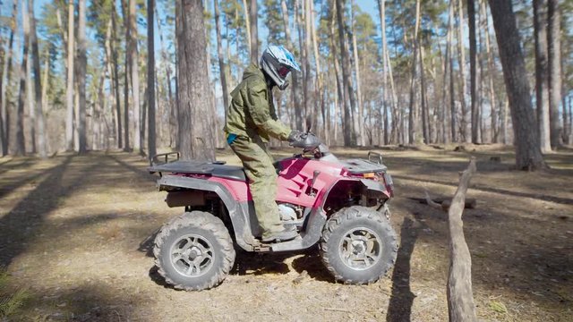 A man in a protective helmet who rides on quad bike in the autumn forest crosses small barriers in the form of trees and branches that are on the road