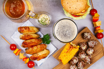 Glasses of beer with chicken wings, burger, meat balls, grilled corn and vegetables. Beer bites. Ale and food still life. View from above, top studio shot