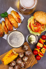 Glasses of beer with chicken wings, burger, meat balls, grilled corn and vegetables. Beer bites. Ale and food still life. View from above, top studio shot