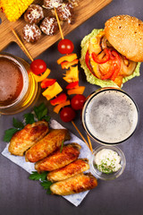 Glasses of beer with chicken wings, burger, meat balls, grilled corn and vegetables. Beer bites. Ale and food still life. View from above, top studio shot