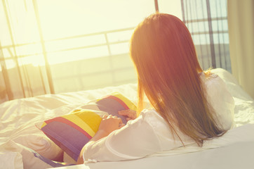 Young asian woman in pajamas sitting and relaxing on the bed while looking at sunlight at window