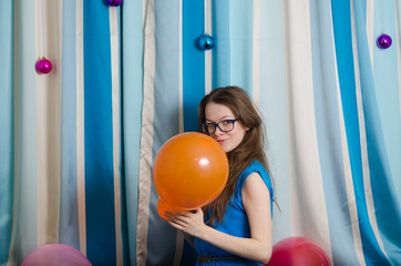 A young woman with glasses and loose hair is playing with balloons. Dressed in a blue dress on a light background. Emotion is playfulness.