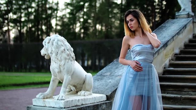 Attractive girl in silver and blue dress stands on stairs with stone balustrade near lion statue posing with hand on neck and turns to the left during photo shoot in antique estate.
