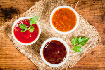 Three kinds of red tomato sauce on a wooden cutting board: traditional classic ketchup, barbecue sauce, sweet and sour Chinese sauce. Top view copy space