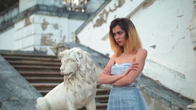 Attractive girl in silver and blue dress stands on stairs with stone balustrade near lion statue posing with arms across and puts arms on waist during photo shoot in antique estate.