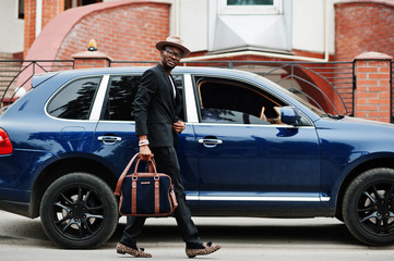 Stylish black man at glasses with hat, wear on suit with handbag against luxury car. Rich african...