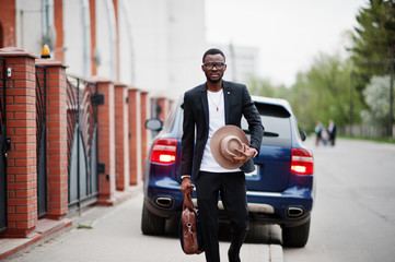 Stylish black man at glasses with hat, wear on suit with handbag against luxury car. Rich african american businessman.