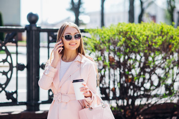Fototapeta premium Upper class woman . Fashionable woman texting outdoors. Fashion woman in a sunglasses and pink jacket with coffee