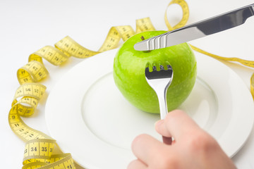Diet concept, green apple on a white plate, yellow measuring tape, fork, knife, woman cuts fruit,...