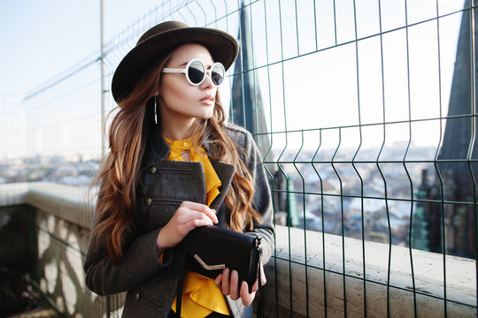 Outdoor waist up portrait of young beautiful woman with long hair, looking aside. Model wearing stylish hat,  white round sunglasses, clothes, holding small bag. City lifestyle. Female fashion concept