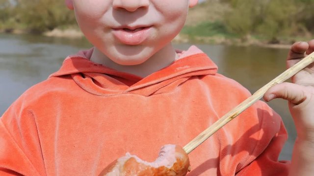 The kid eats the sausage with his teeth, strung on the willow twig with pleasure.
