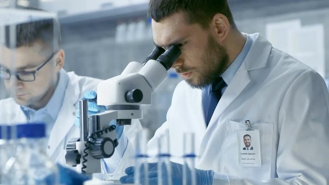 In a Modern Laboratory Two Scientists Conduct Experiments. Chief Research Scientist Dictates the Results He Sees in a Microscope to His Assistant. Shot on RED EPIC-W 8K Helium Cinema Camera.