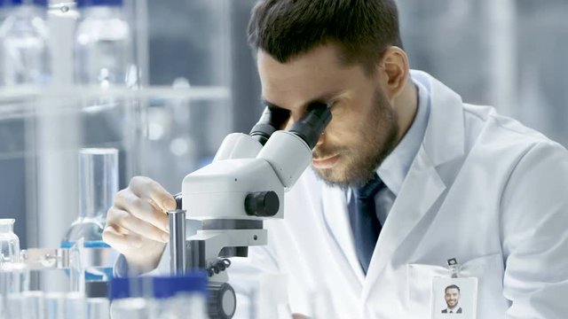 Research Scientist Adjusts His Microscope. He's Working in a High-End Modern Laboratory with Beakers, Glassware, Microscope and Working Monitors Surround Him. 