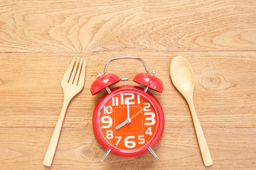 Red alarm clock in wooden dish, spoon and fork on wooden plank background. Time eating concept