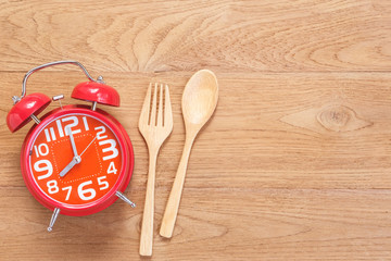Red alarm clock in wooden dish, spoon and fork on wooden plank background. Time eating concept