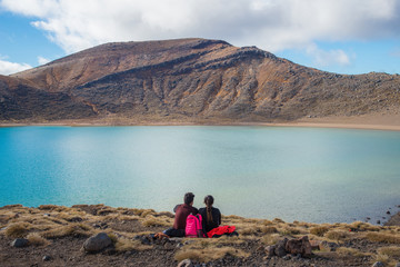 Fototapeta premium Couple sitting and relax with the spectacular view of Blue lake during trekking in Tongariro Alpine Crossing in New Zealand.