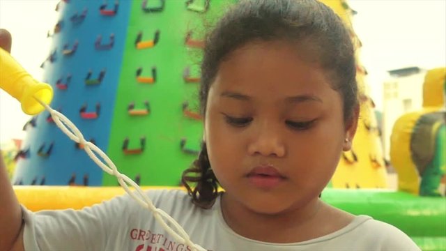 Close up of a multicultural Asian girl blowing bubbles with an inflatable climbing mountain behind her (100fps clow motion)