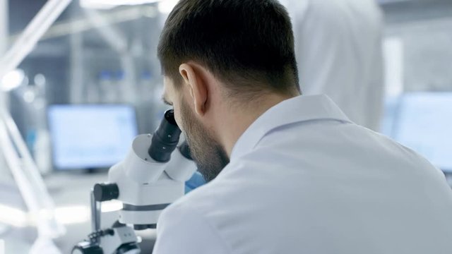 Over the Shoulder View of a Research Scientist Looking into Microscope. He's Conducts Experiments with His Colleagues in Modern Laboratory. Shot on RED EPIC-W 8K Helium Cinema Camera.