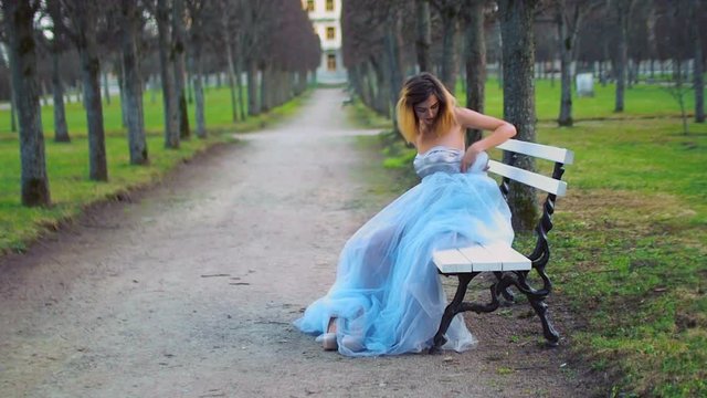 Attractive girl in silver and blue dress sits on bench in parkway adjusting dress and getting ready to pose during photo shoot.
