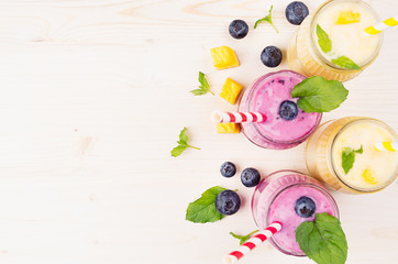 Naklejka na ściany i meble Freshly blended yellow and violet fruit smoothie in glass jars with straw, mint leaves, mango slices and berry, top view. Soft white wooden board background, copy space.
