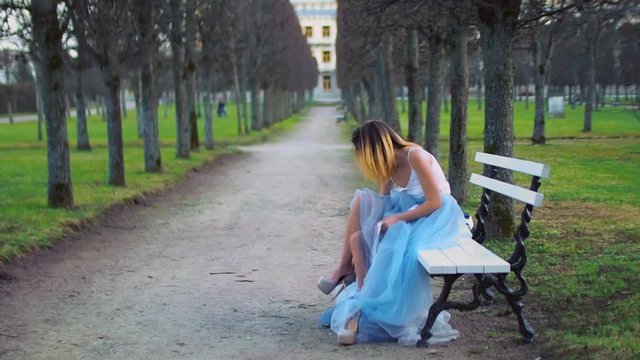 Attractive girl in silver and blue dress sits on bench in parkway putting on high heeled shoes and gets ready to pose during photo shoot. Side view.