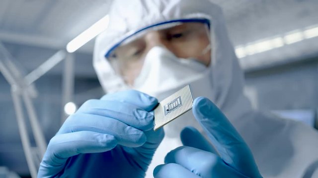 In Ultra Modern Electronic Manufacturing Factory Design Engineer in Sterile Coverall Holds Microchip with Gloves and Examines it. Shot on RED EPIC-W 8K Helium Cinema Camera.