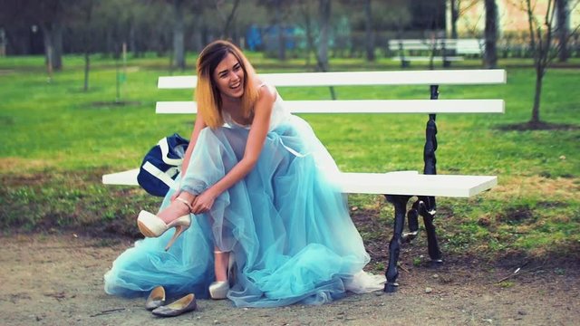 Attractive girl in silver and blue dress sits on bench in parkway putting on high heeled shoes, poses and stands up from bench during photo shoot. Front view.