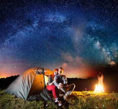 Night camping. Charming couple tourists sitting in front tent near campfire under shines starry sky and Milky way