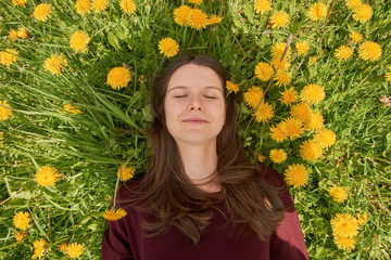 Smiling young woman with closed eyes relaxing on a meadow with many dandelions in the spring sun. Top down view.