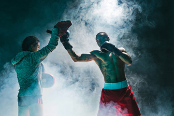 Handsome Afro American boxer with bare torso is practicing punches with a partner at the fight club