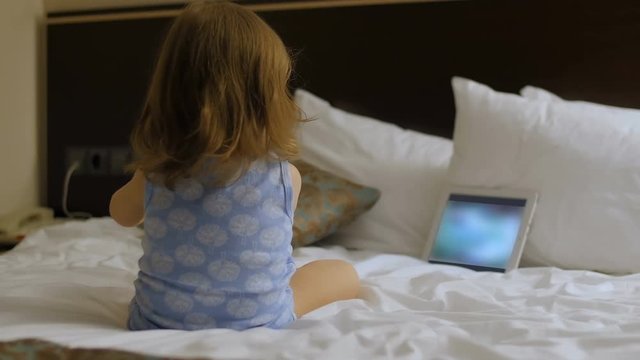 A child is sitting in the bedroom on the bed looking at the phone, tablet, computer.