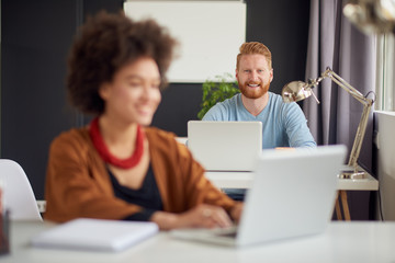 Young Caucasian businessman sitting and using laptop computer in modern office. In background colleague