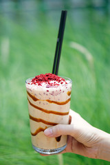 Salty Caramel Shake (Blended Coffee) with Red Velvet Crumbles, Hand holding glass of Coffee frappe (frappuccino) with caramel syrup, whipped cream and crumble on grassland background.