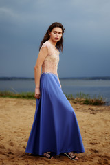 Fototapeta na wymiar Calm lonely fashion model walking on the sand in a cloudy day. Romantic, gentle, mystical, image of a girl in long blue skirt and lace blouse.